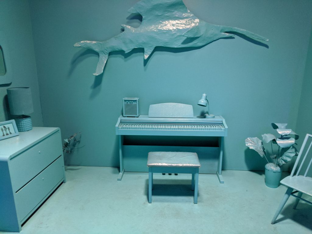 A bedroom in which everything is painted the same color turquoise. There is a fish, a piano, a dresser, and some other items in the room.