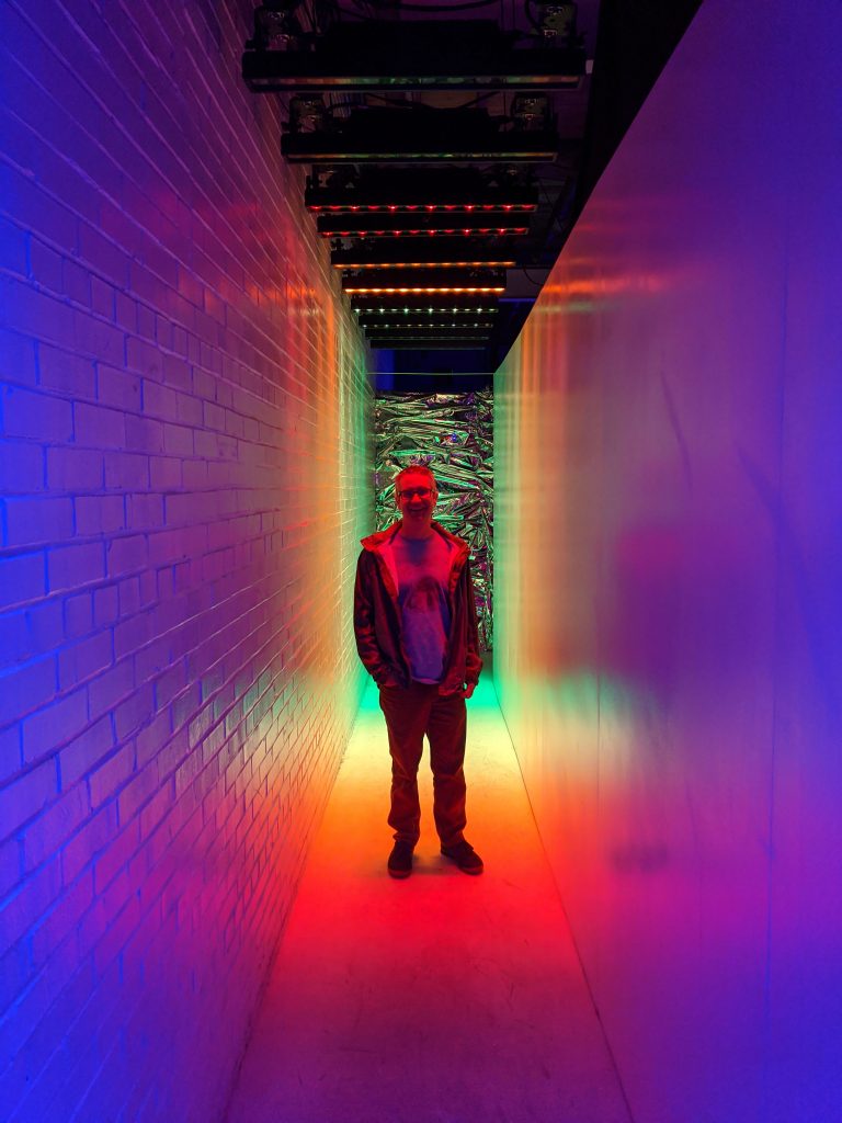 A narrow space filled with a rainbow of color. A man stands in the middle looking at the camera.
