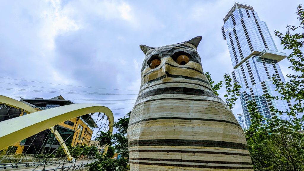 A unique owl statue gazes at the camera, with a bridge and skyscraper in the background.