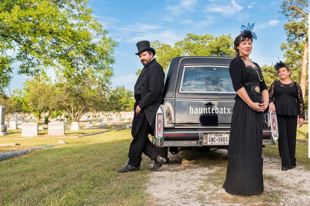 Three people in black mourning clothing standing next to a hearse.
