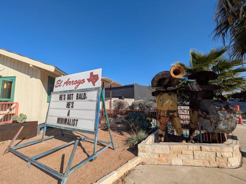The El Arroyo sign reading "He's not bald, he's a minimalist" next to rusty metal mariachi sculptures.