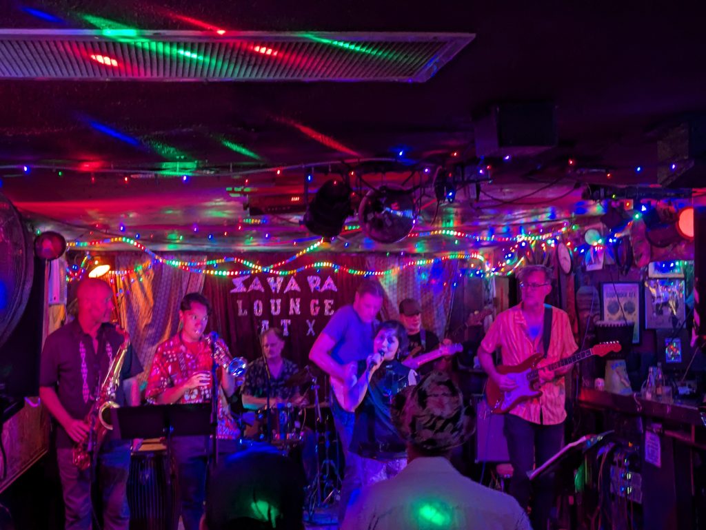A band with saxophone, trumpet and guitars playing under string lights in front of the Sahara Lounge ATX backdrop.